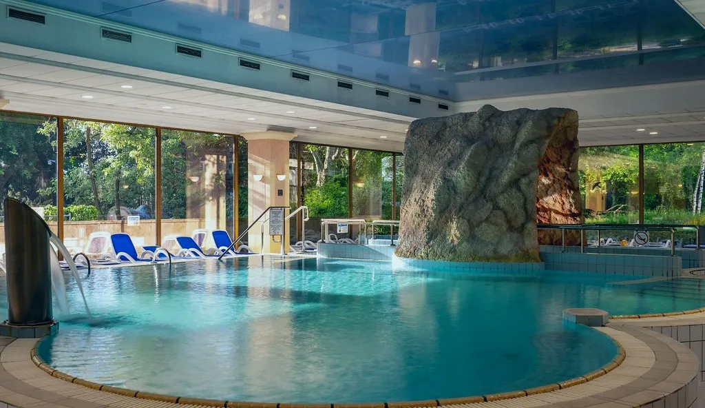 Az Ensana Thermal Margitsziget Health Spa beltéri termálvizes medencéje, ahol szikla dekoráció, türkiz nyugágyak és panorámaablakok láthatók