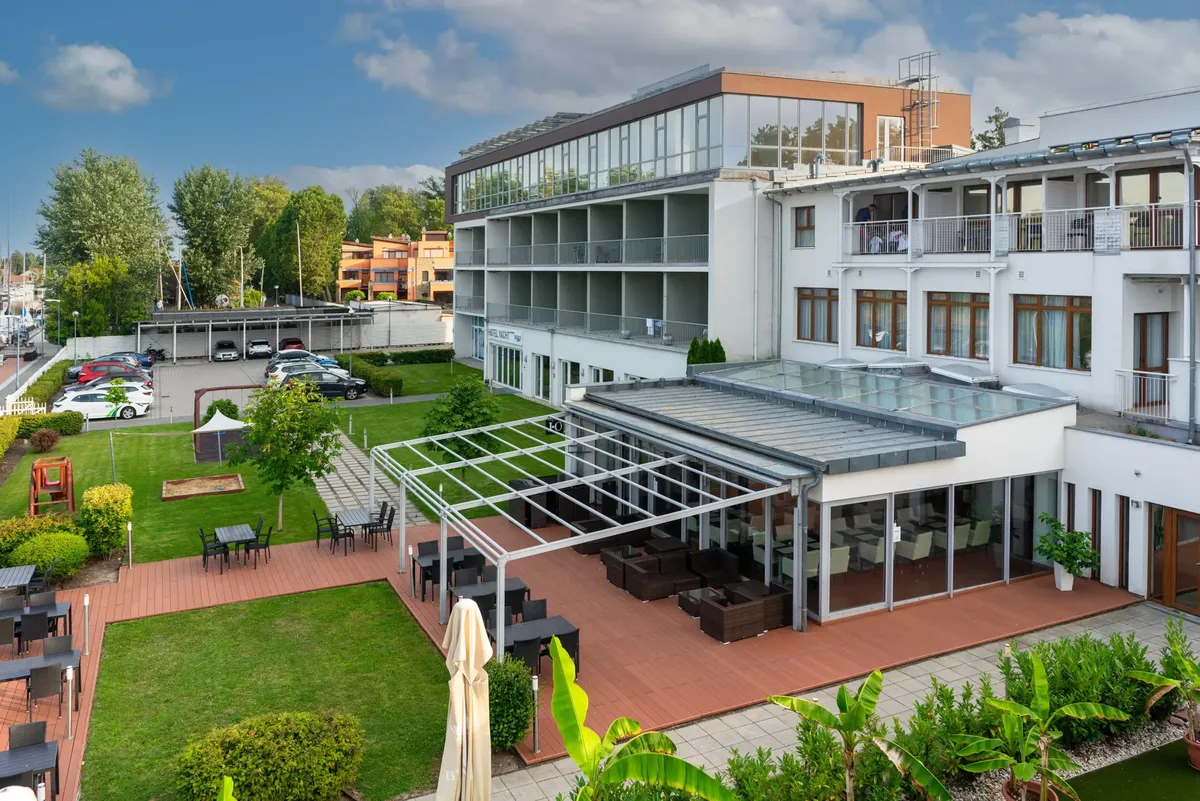 A Hotel Yacht Siófok parkosított kertje és teraszai, ahol közösségi terek, üvegezett étterem és modern épület látható