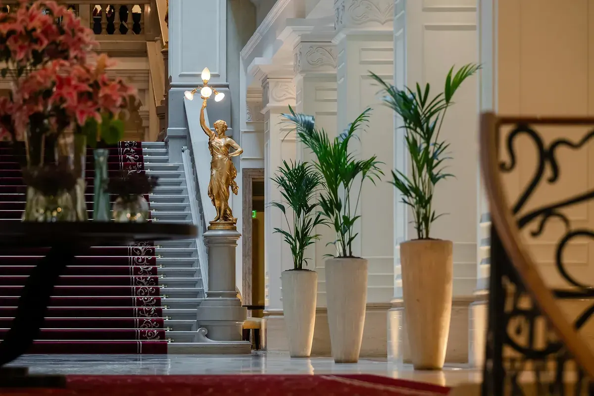 Corinthia Budapest főlépcső, arany szobor, neoklasszikus oszlopok, bordó futószőnyeg, luxus hotel